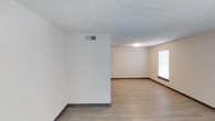 This single-floor space offers a straightforward and practical layout designed for comfortable everyday living. The interior features light wood-style laminate flooring throughout, which lends warmth and continuity to the open areas. Neutral, light-toned walls create a bright and airy atmosphere, while a textured ceiling adds a subtle layer of visual detail overhead. Dark baseboards provide a clean contrast against the pale walls and flooring, giving the space a polished, finished look. A ceiling-mounted light fixture provides consistent overhead illumination, complementing the natural light that filters in through a window fitted with horizontal blinds. The HVAC vent positioned on the upper wall indicates a central air system, contributing to year-round comfort. The open floor plan allows for flexible furniture arrangements and accommodates a range of living configurations. The overall ambiance is clean, minimal, and move-in ready, making it a versatile space well-suited for those seeking a simple, functional environment with a neutral palette as a backdrop for personalization.