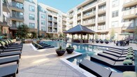 This pool area is designed as a comprehensive outdoor retreat, combining multiple functional zones within a well-appointed courtyard setting. At the heart of the space is a substantial wood-framed cabana with a pitched metal roof and exposed timber beam ceiling, providing shaded seating with plush, cushioned sofas accented by green throw pillows—oriented to overlook the sparkling pool. The expansive pool itself features a central water fountain feature and is flanked by generous sun decks lined with chaise lounges, offering ample space for relaxation.

An outdoor kitchen and bar area serves as a central amenity hub, constructed with brick detailing and stainless steel appliances, a built-in sink, and a wall-mounted flat-screen TV. Modern pendant lighting and white cabinetry complete the polished look. Wicker bar seating surrounds white countertops, creating a social gathering space with a warm wood-paneled ceiling overhead.

Additional covered lounge areas feature wicker armchairs, side tables, and bistro-style seating arrangements set on tiled pavers. Lush container plantings and mature trees are thoughtfully integrated throughout, enhancing the resort ambiance. The entire area is framed by the surrounding multi-story building, creating an intimate and private outdoor environment.