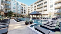 This pool area is designed as a comprehensive outdoor retreat, combining multiple functional zones within a well-appointed courtyard setting. At the heart of the space is a substantial wood-framed cabana with a pitched metal roof and exposed timber beam ceiling, providing shaded seating with plush, cushioned sofas accented by green throw pillows—oriented to overlook the sparkling pool. The expansive pool itself features a central water fountain feature and is flanked by generous sun decks lined with chaise lounges, offering ample space for relaxation.

An outdoor kitchen and bar area serves as a central amenity hub, constructed with brick detailing and stainless steel appliances, a built-in sink, and a wall-mounted flat-screen TV. Modern pendant lighting and white cabinetry complete the polished look. Wicker bar seating surrounds white countertops, creating a social gathering space with a warm wood-paneled ceiling overhead.

Additional covered lounge areas feature wicker armchairs, side tables, and bistro-style seating arrangements set on tiled pavers. Lush container plantings and mature trees are thoughtfully integrated throughout, enhancing the resort ambiance. The entire area is framed by the surrounding multi-story building, creating an intimate and private outdoor environment.