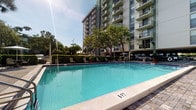 This outdoor amenity area centers around a generously sized swimming pool with crystal-clear turquoise water, reaching a depth of six feet at its far end. The pool is framed by a clean, light-toned tile surround that provides ample space for poolside relaxation. A row of lounge chairs lines the perimeter, offering residents a comfortable place to unwind in the sun or shade. Mature trees and well-maintained hedges create a natural privacy buffer, adding a touch of greenery to the setting. The surrounding landscape is neatly manicured, contributing to an overall polished and welcoming atmosphere. In the background, a tall multi-story building rises prominently, with balconies on each floor overlooking the pool area. Additional covered parking and shade structures are visible nearby. The open-sky setting allows for abundant natural sunlight throughout the day, enhancing the bright and airy feel of the space. Overall, this outdoor area presents a well-appointed communal retreat designed for leisure and recreation.