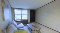 This single-floor space features a living area designed with simplicity and functionality in mind. The room is finished with light-toned walls accented by classic crown molding, lending a clean and structured appearance to the interior. Neutral carpeting runs throughout, contributing to a warm and understated aesthetic.

A large window fitted with vertical blinds serves as the room's primary source of natural light, allowing outdoor views while maintaining privacy control. The window is flanked by a wood-paneled base, adding a subtle rustic contrast to the otherwise soft palette. A bold, dark-stained six-panel entry door anchors one wall, providing a strong visual focal point against the lighter surroundings.

The living area accommodates a light grey sofa with accent cushions and a rustic wooden coffee table with a lower shelf, offering both comfort and practical storage. A patterned area rug grounds the seating arrangement, adding texture to the carpeted floor. The overall ambiance of the space is calm and neutral, with an approachable, lived-in character suited for everyday comfort.