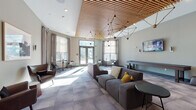 This thoughtfully designed single-floor amenity space combines contemporary aesthetics with multifunctional areas to create a dynamic and inviting environment. The centerpiece is a spacious lounge area distinguished by soaring ceilings adorned with warm wood slat panels and striking geometric pendant light fixtures that add sculptural character to the space. Large floor-to-ceiling windows flood the room with natural light, while plush gray sectional sofas, accent chairs, and small bistro tables provide versatile seating arrangements suited for both socializing and relaxation. A shuffleboard table and wall-mounted flat-screen television anchor the entertainment zone, complementing the lounge's recreational appeal. Adjacent to the lounge, a well-appointed kitchenette features a dark-toned island with bar seating, cabinetry, and a sink, all set against rich hardwood flooring that transitions seamlessly through the space. Glass-walled private offices or conference rooms line one side of the floor, offering transparency and accessibility while maintaining functional separation. The overall design palette of soft grays, warm wood tones, and muted greens creates a cohesive, modern ambiance that balances comfort with sophistication throughout every area of the space.