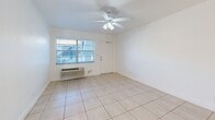 Located at 1860 SW 68th Ave in Miramar, FL, this one-bedroom, one-bathroom unit offers a compact and efficiently designed single-floor layout. Light beige ceramic tile flooring runs consistently throughout every room, lending a clean, cohesive look to the entire space. The living room is a bright, open area featuring a ceiling fan with light fixture, a wall-mounted air conditioning unit, and a window fitted with horizontal blinds that allows natural light to filter in. The space flows openly toward the kitchen, which is a standout feature of the unit, showcasing white cabinetry, granite-look countertops, a decorative mosaic tile backsplash, and stainless steel appliances including a range, microwave, and additional natural light from a window above the counter area. The bedroom is a generously proportioned space that includes a ceiling fan, a sliding double-door closet, a wall AC unit, and a window overlooking the exterior. The bathroom is finished with full white ceramic wall tile surrounding a shower/tub combination, complemented by a window that provides natural ventilation and light. Throughout the unit, crisp white walls and consistent flooring create a bright, neutral palette well-suited for a variety of interior styles.