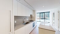 This thoughtfully designed one-bedroom, one-bathroom unit offers a clean, contemporary aesthetic across a single floor. The open-concept living and kitchen area is anchored by a streamlined galley-style kitchen, featuring flat-front white cabinetry extending to the ceiling, a taupe quartz backsplash and countertops, a stainless steel undermount sink, gas cooktop, and built-in oven. A central kitchen island provides additional prep and dining space. Light hardwood flooring runs consistently throughout, enhancing the bright, airy atmosphere.

The living area is defined by expansive floor-to-ceiling windows with black frames and roller shades, offering wide urban views of the surrounding cityscape and filling the space with abundant natural light. The bedroom is well-proportioned, with the same large-format windows framing outdoor views, and features a sliding closet and access to additional closet storage.

The bathroom is finished to a high standard, with large-format grey tiles, a frameless glass walk-in shower, a floating white vanity with an integrated sink, and a wide mirror. A separate walk-in closet area with built-in shelving adjoins the bathroom. Recessed lighting throughout adds a polished, modern touch to this well-appointed urban residence.