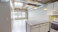 This two-floor loft showcases a striking industrial aesthetic that blends raw architectural character with modern finishes. The space is distributed across two levels, with the lower floor housing the main living and kitchen areas, and the upper floor providing additional living space.

The kitchen is a standout feature, equipped with granite countertop surfaces, a large central island, white cabinetry, stainless steel appliances including a range, microwave, and dishwasher, and a vibrant yellow subway tile backsplash that adds a bold pop of color. Pendant lights hang from the exposed concrete beam ceiling, which also features industrial-style ductwork running overhead — a defining characteristic of the space's loft-style identity.

Large multi-pane windows allow generous natural light to flood the open living area, offering views of the surrounding exterior. Polished concrete flooring anchors the lower level, contributing to the space's urban, warehouse-inspired feel.

The walk-in closet is outfitted with adjustable wire shelving and hanging rods, providing versatile and well-organized storage. The bathroom features a combination tub and shower, tile flooring, and yellow accent tiling that echoes the kitchen's design palette, creating a cohesive visual theme throughout. A hallway connects the key areas of the space, contributing to a practical and efficient layout across both floors.