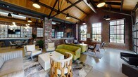 This single-story space showcases a striking industrial-chic design, seamlessly blending raw architectural character with modern, curated furnishings. The most commanding feature is the soaring vaulted ceiling, finished with warm wood planking and reinforced by bold black steel trusses, lending the space impressive vertical drama. Exposed brick walls run throughout, complemented by polished concrete floors that reflect the abundant natural light pouring in through oversized steel-framed windows and a full-panel glass garage door that opens to an outdoor courtyard beyond.

The open floor plan is thoughtfully organized into distinct functional zones. A lounge area anchors one section, furnished with dark leather sofas and accent chairs arranged around a low coffee table. Adjacent seating vignettes feature eclectic mid-century and contemporary furniture, including a moss-green tufted sofa and sculptural wood accent pieces. A bar and counter area with barstool seating provides a social hub, while a dining zone with wood tables and upholstered chairs offers flexible gathering space.

A media wall with a mounted flat-screen television and a decorative display installation serves as a focal point near the entrance. Pendant lighting clusters, industrial drop lights, and globe fixtures layer warm illumination throughout. The overall ambiance is energetic yet comfortable, ideal for a communal or event-oriented setting.