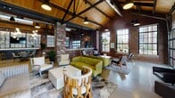This single-story space showcases a striking industrial-chic design, seamlessly blending raw architectural character with modern, curated furnishings. The most commanding feature is the soaring vaulted ceiling, finished with warm wood planking and reinforced by bold black steel trusses, lending the space impressive vertical drama. Exposed brick walls run throughout, complemented by polished concrete floors that reflect the abundant natural light pouring in through oversized steel-framed windows and a full-panel glass garage door that opens to an outdoor courtyard beyond.

The open floor plan is thoughtfully organized into distinct functional zones. A lounge area anchors one section, furnished with dark leather sofas and accent chairs arranged around a low coffee table. Adjacent seating vignettes feature eclectic mid-century and contemporary furniture, including a moss-green tufted sofa and sculptural wood accent pieces. A bar and counter area with barstool seating provides a social hub, while a dining zone with wood tables and upholstered chairs offers flexible gathering space.

A media wall with a mounted flat-screen television and a decorative display installation serves as a focal point near the entrance. Pendant lighting clusters, industrial drop lights, and globe fixtures layer warm illumination throughout. The overall ambiance is energetic yet comfortable, ideal for a communal or event-oriented setting.