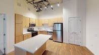 This single-floor loft-style unit showcases an industrial-chic aesthetic defined by soaring ceilings with exposed concrete soffits, visible steel ductwork, and track lighting throughout. The open living area is anchored by rich, dark hardwood flooring that reflects abundant natural light streaming in through large windows fitted with vertical blinds. The spacious layout flows seamlessly from the living area into a well-designed kitchen, which features light maple cabinetry extending to the ceiling, stainless steel appliances including a refrigerator, range, microwave, and dishwasher, white countertops, and a central island that provides additional prep and dining space. Light wood flooring in the kitchen complements the warm cabinetry tones. The bedroom offers plush carpeting, a large window with outdoor views, and the same signature exposed concrete ceiling detail with ductwork, reinforcing the industrial character of the space. A hallway connects the main living areas and provides access to the bathroom, which is outfitted with a vanity featuring maple cabinetry, a white countertop, a large mirror, hardwood-style flooring, and a tub-shower combination. A stacked washer and dryer unit is conveniently housed within the bathroom area, adding practical functionality to this distinctively styled urban residence.