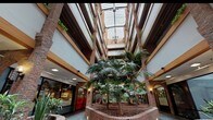 The Main Level Atrium is an architecturally distinctive indoor space that rises dramatically through multiple stories, culminating in a large skylight that floods the interior with natural light. Exposed brick columns anchor the design, complementing warm wood beam accents and white-paneled upper balconies that line the perimeter of each floor above. Lush tropical palm trees grow from generously sized raised brick planters at the center of the atrium, creating a verdant, garden-like atmosphere. Trailing greenery cascades from the upper-level railings, enhancing the organic character of the space. The main level floor is finished in a distinctive teal-toned square tile that adds color and texture underfoot, while curved brick planter walls serve as informal seating areas with wooden bench surrounds. A glass elevator shaft rises at the heart of the atrium, providing both functional access to upper floors and a sleek architectural focal point. Retail and commercial storefronts line the perimeter, their large glass facades opening onto the walkways. Wall-mounted lantern-style sconces and overhead track lighting provide warm, even illumination throughout. The overall ambiance is welcoming and lively, blending natural elements with classic brick-and-timber architecture to create an inviting communal environment.