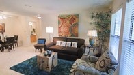 This single-floor space offers a comfortable and thoughtfully arranged living area that exudes a welcoming ambiance. The living room serves as the centerpiece, featuring a rich, dark upholstered sofa adorned with decorative "HOME" pillows, complemented by a patterned accent chair that adds a playful, eclectic touch. A bold, colorful canvas artwork anchors the main wall, infusing the room with vibrant energy through its warm reds, teals, and earthy tones. The space is grounded by a deep teal area rug that adds depth and color contrast to the neutral, light-toned walls and flooring. Soft, layered lighting from a floor lamp and a table lamp with a decorative ceramic base creates a warm, inviting glow throughout the room. A unique side table with Parisian-inspired print detailing serves as a conversation piece alongside a wrought-iron accent table. Lush indoor bamboo-style plants in the corner bring a natural, organic element to the interior. An open sightline leads into an adjacent bathroom area, hinting at a functional and connected floor plan. Overall, the space balances bold decorative choices with a cozy, lived-in comfort.
