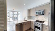 This single-floor space showcases a thoughtfully designed, modern interior with an emphasis on clean lines and high-end finishes. The open-concept kitchen serves as a focal point, outfitted with premium stainless steel appliances including a professional-grade gas range and built-in microwave. A peninsula with a crisp white quartz countertop provides additional prep space and casual seating, complemented by an undermount sink with a sleek modern faucet. The cabinetry features a contemporary flat-front design in a warm taupe wood-grain finish, offering generous storage throughout. A textured stone mosaic tile backsplash adds visual depth and contrast against the light countertops. Hardwood flooring anchors the kitchen area, transitioning to a polished hard surface in the adjacent living space. Floor-to-ceiling glass doors and windows flood the interior with natural light and connect seamlessly to an outdoor area, creating an open and airy ambiance. The high white ceilings with recessed lighting further enhance the sense of spaciousness, making this a refined and functional modern living environment.