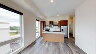 This contemporary single-floor residence showcases a thoughtfully designed open-concept layout with clean lines and modern finishes throughout. The kitchen serves as a central highlight, featuring warm wood cabinetry with glass-front upper panels, a built-in wine rack, white quartz countertops, a subway tile backsplash, and a full suite of stainless steel appliances including a range with hood ventilation, dishwasher, and a French door refrigerator with ice dispenser. A sleek kitchen island with a matching wood veneer base and white quartz surface provides additional prep and dining space, illuminated by elegant glass pendant lights overhead.

The living area is generously proportioned, bathed in natural light through large floor-to-ceiling windows and a sliding glass door that opens to a private balcony overlooking the surrounding urban landscape. Hardwood-style flooring flows throughout the main living spaces, while the bedrooms feature plush carpeting and large windows that frame views of the cityscape.

The primary bathroom is well-appointed with a floating double vanity, backlit mirrors, wood cabinetry, and a tiled walk-in shower. A secondary bathroom includes a soaking tub and hardwood-style flooring. The overall ambiance is bright, contemporary, and well-suited for modern urban living.