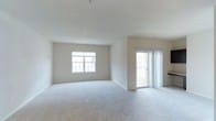 This single-floor space presents a clean, open layout finished in a neutral, light beige palette that contributes to a calm and airy atmosphere throughout. The main living area is generously proportioned, featuring wall-to-wall plush carpeting in a soft gray-beige tone that adds warmth underfoot while complementing the overall color scheme. Smooth white ceilings with recessed lighting provide even, ambient illumination across the room.

Two sources of natural light define the space: a multi-pane window centered on the far wall invites daylight deep into the interior, while a sliding glass door flanked by sheer white curtains offers access to an outdoor area, likely a balcony or patio. The combination of these openings creates a bright, connected feel between the indoors and outdoors.

The walls are finished in a clean, off-white tone, providing a neutral backdrop suitable for a variety of furnishing styles. The layout is open and unobstructed, allowing for flexible furniture arrangement. Overall, the space offers a move-in-ready feel with a simple, functional design well-suited to comfortable everyday living.