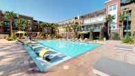 This expansive outdoor amenity space is designed with a resort-inspired aesthetic, offering a comprehensive array of leisure and recreation features set against a backdrop of towering palm trees and a multi-story contemporary building facade. At the heart of the space lies a generously sized swimming pool with sparkling turquoise water, complemented by a separate spa, paver pool decks lined with chaise lounges, and shaded umbrella seating areas for sunbathing and relaxation. A covered outdoor lounge area provides a comfortable retreat, equipped with a ceiling fan, a modern linear gas fireplace, plush sectional seating in cool grey tones accented with green pillows, and a foosball table for entertainment. Adjacent to the pool area, dual stainless steel built-in BBQ grills are set into a dedicated outdoor kitchen counter, ideal for gatherings. A striking circular hammock structure with a canopy shade offers a unique relaxation spot surrounded by lush landscaping. A sculptural woven rattan cabana features a full wraparound cushioned sectional with green upholstery, creating an intimate outdoor lounge pod. An additional covered cabana room includes casual seating, a high-top table, ceiling fan, and decorative wall art, providing a shaded, semi-private gathering space.