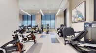 This amenity floor is defined by two distinct yet complementary spaces: a sophisticated lobby lounge and a comprehensive fitness center. The lobby makes an immediate impression with its soaring ceilings and a dramatic recessed oval ceiling detail housing a striking crystal chandelier. Large floor-to-ceiling windows framed in dark steel flood the space with natural light, while large-format light stone tile flooring anchors the open layout. A bold botanical wall art installation serves as a focal point, complemented by a dark wood feature wall with an integrated flat-screen display. Comfortable seating arrangements with plush velvet chairs and textured area rugs define conversation areas, creating an inviting, hotel-caliber atmosphere. A staircase and elevator bank with textured wave-pattern wall tile provide polished vertical circulation.

The fitness center is spacious and well-appointed, organized into distinct cardio and strength training zones. The cardio area features multiple treadmills, ellipticals, upright and recumbent bikes, and a rowing machine, all positioned to take advantage of large exterior-facing windows with outdoor views. A dedicated strength area includes a cable machine, a full dumbbell rack, kettlebells, a flat bench, and wall-mounted mirrors and television, rounding out a thoroughly equipped workout facility.