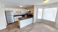 This single-floor space showcases a thoughtfully designed open-concept layout that seamlessly connects the kitchen and living areas. The kitchen is a standout feature, boasting crisp white cabinetry, dark granite-style countertops, and stainless steel appliances, including a refrigerator and electric range. A peninsula with matching countertops extends the workspace and provides an ideal spot for casual dining or entertaining. Pendant lighting above the peninsula adds a stylish focal point, complementing the flush-mount ceiling fixtures throughout. The kitchen also includes a stainless steel sink set beneath upper cabinetry, with a range hood tucked neatly above the stove. Wood-look flooring runs through the kitchen area, transitioning to plush carpeting in the adjacent living space, creating a warm and inviting atmosphere. A large bay-style window floods the living area with natural light, enhancing the open and airy feel of the space. Neutral wall tones and a textured ceiling tie the design together, offering a clean, contemporary aesthetic throughout. The overall layout is functional and well-appointed, making excellent use of the available space.
