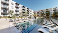 Located at 305 W Commerce St in Dallas, Texas, this outdoor amenity space is designed to deliver a resort-style experience within a modern residential community. The centerpiece is a large, beautifully maintained swimming pool with crystal-clear water, flanked on both sides by generous sun decks lined with contemporary lounge chairs and chaise lounges, ideal for relaxing under the Texas sun. A cascading water feature adds a dynamic visual element along one edge of the pool, enhancing the overall ambiance. Adjacent to the pool, a covered outdoor pavilion provides a shaded gathering area equipped with ceiling fans, mounted flat-screen televisions, and ample seating at large communal tables — perfect for outdoor dining or social events. The surrounding landscape incorporates ornamental grasses, trees, and manicured greenery that soften the hardscape and create an inviting atmosphere. A dedicated outdoor grilling station with a stainless steel built-in grill offers a convenient space for cookouts. The entire amenity area is framed by the multi-story residential building, featuring private balconies that overlook the courtyard. The space strikes a balance between active recreation and relaxed outdoor living, making it a well-appointed communal environment in the heart of Dallas.