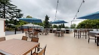 This expansive outdoor patio serves as a well-appointed communal gathering space, thoughtfully designed to accommodate a variety of social activities. The open-air layout features a smooth, light-toned concrete deck that provides ample room for movement and socializing throughout the area.

The patio is divided into distinct functional zones. A central BBQ section includes multiple built-in grills set into sleek concrete stations, flanked by high-top tables and bar-height seating — ideal for outdoor dining and entertaining. Several large blue market umbrellas are positioned throughout to provide shade, complementing the warm-toned wood and metal outdoor furniture.

A covered portion of the patio, adjacent to the clubhouse building, offers a sheltered lounge area with comfortable navy-cushioned armchairs and coffee tables, perfect for relaxed conversation. The clubhouse itself features classic gray shingle siding, white trim, and a warm wood-paneled ceiling overhang, contributing a coastal, residential aesthetic. Large windows along the building facade allow views between the interior and exterior spaces.

Overhead string lights are strung across the open deck, creating an inviting ambiance for evening gatherings. The surrounding landscape includes mature trees and views of neighboring residential buildings, providing a lush, community-oriented backdrop throughout the space.