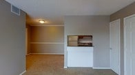This single-floor space offers a straightforward and functional layout designed for comfortable everyday living. The main living area is finished with wall-to-wall carpeting in a warm, neutral tone, creating a cozy and cohesive feel throughout. The walls are painted in a soft gray hue, contributing to a calm and understated ambiance.

A defining architectural feature is the pass-through kitchen window, which opens directly from the kitchen into the living area. This design element promotes an open, connected feel between the two spaces while maintaining a clear spatial distinction. The kitchen, visible through the pass-through, includes cabinetry with a warm wood finish, a refrigerator, and a sink with countertop space, offering essential functionality in a compact footprint.

The living area benefits from a textured ceiling with flush-mount lighting, providing even illumination throughout the space. A subtle white chair rail runs along the perimeter of the living area walls, adding a modest decorative detail that lends a touch of classic character to the otherwise minimalist interior. White baseboards complete the trim work, providing clean visual boundaries at floor level. Overall, the space presents a clean, neutral, and practical environment well-suited for a variety of uses.