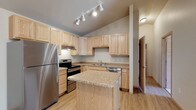 This single-floor residence offers a thoughtfully designed layout with a variety of functional living spaces. The kitchen serves as a standout feature, showcasing light wood cabinetry that extends to the upper walls, granite countertops, and a central island that provides additional prep space and casual seating. Stainless steel appliances — including a refrigerator, electric range, and dishwasher — complement the warm, neutral tones throughout. Track lighting overhead ensures the kitchen is well-illuminated and practical. Hardwood-style flooring flows throughout the main living and kitchen areas, lending a cohesive and warm aesthetic to the space. The floor plan reveals a generously sized open area in the lower half of the layout, offering flexible use as living and dining zones. A full bathroom is visible in the upper right of the floor plan, complete with a soaking tub and additional fixtures. A dedicated hallway connects the various rooms, providing clear circulation throughout. The overall ambiance is bright and inviting, with a clean, modern sensibility anchored by natural wood tones and neutral wall colors that make the space feel open and welcoming.