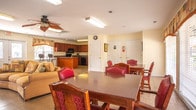 Located at 4441 E Honeygrove Ct in Virginia Beach, Virginia, this single-story space offers a welcoming and functional layout designed for comfortable communal use. The open-plan interior is unified by light-toned tile flooring that flows seamlessly throughout, creating a cohesive and easy-to-navigate environment.

The spacious living area is a central gathering point, furnished with a large sectional sofa, leather accent chairs, and a substantial wood entertainment center housing a television and built-in shelving. A decorative ceiling fan adds to the comfortable ambiance, while natural light filters in through windows dressed with patterned valances.

Adjacent to the living space, the dining area features a large wooden table with upholstered chairs, set beneath arched windows with views of an outdoor patio area. The generous window arrangement allows ample natural light to brighten the room throughout the day.

The kitchen is well-appointed with rich cherry wood cabinetry, dark granite countertops, and black appliances including a range, microwave, dishwasher, and refrigerator. A central island with bar seating provides additional workspace and a casual dining option. Recessed and overhead lighting ensure the kitchen is well-lit for meal preparation. Overall, the space offers a practical and inviting setting suited for group activities and shared dining.