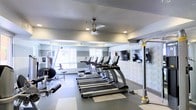 This fitness center offers a clean, modern environment designed to support a well-rounded exercise routine. The space is finished with neutral gray walls accented by a subtle horizontal stripe detail, giving it a contemporary and polished aesthetic. Durable, low-profile flooring covers the workout area, providing both stability and ease of maintenance.

A row of TRU treadmills lines the center of the room, positioned to face a bank of wall-mounted flat-screen televisions, allowing users to stay entertained during cardio sessions. To the left, a multi-function strength training machine provides options for resistance-based workouts. Additional equipment is visible along the perimeter, rounding out the variety of fitness options available.

Large windows along the far wall welcome an abundance of natural light, creating an open and energizing atmosphere. A ceiling fan with integrated lighting is centrally mounted overhead, promoting air circulation and comfort during workouts. Recessed lighting supplements the natural light, ensuring the space remains well-lit at all hours. Overall, the fitness center balances functionality with a bright, inviting ambiance suited for daily use.