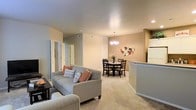 This single-floor residence offers a thoughtfully designed open-concept layout that seamlessly connects the living, dining, and kitchen areas into one cohesive and inviting space. The living area is anchored by a comfortable sofa accented with warm-toned throw pillows, complemented by plush wall-to-wall carpeting that adds a cozy feel underfoot. Neutral beige walls create a warm, versatile backdrop throughout the space.

The dining area is positioned adjacent to the living room and features a classic dark wood dining table with matching chairs, illuminated by an elegant chandelier that adds a refined touch to the room. A decorative wall arrangement near the dining space adds a personalized artistic accent.

The kitchen is partially visible from the main living area, separated by a curved counter or breakfast bar that provides a natural transition between spaces. White cabinetry and appliances contribute to a clean, bright aesthetic in the kitchen zone.

Recessed lighting and ceiling-mounted fixtures ensure the space is well-lit throughout, while interior doorways lead to additional rooms, suggesting a practical and functional floor plan. Overall, the residence exudes a warm, comfortable ambiance with a balance of casual and refined design elements.