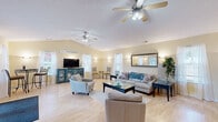 Located at 626 Chelsea Pl in Newport News, VA, this single-floor space offers a thoughtfully designed layout that balances comfort and functionality. The spacious living room is the heart of the space, characterized by vaulted ceilings, warm yellow-toned walls, and light hardwood flooring that enhances the airy atmosphere. Dual ceiling fans provide comfort throughout, while multiple windows dressed with sheer white curtains invite abundant natural light. A decorative arched mirror and wall sconces add elegant touches, complementing the neutral, inviting furnishings. French doors with glass panes open to an outdoor area, further extending the sense of openness. A bistro-style seating area near the entrance offers a casual gathering spot. The connecting hallway leads to a well-equipped exercise room featuring a dark rubberized floor, wall-to-wall mirrors, and a variety of fitness equipment including a treadmill, stationary bike, and multi-use weight machine, illuminated by bright overhead panel lighting. A water cooler adds a practical amenity to the workout space. Together, these rooms create a versatile, welcoming environment suited for both relaxation and an active lifestyle.