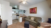 This single-floor unit offers a practical and well-organized open-concept layout, where the living, dining, and kitchen areas flow naturally into one another. The living area is anchored by a comfortable sofa and features carpeted flooring, a ceiling fan with a light fixture overhead, and a textured ceiling. A tufted ottoman serves as a versatile centerpiece, and framed wall art adds a touch of warmth and color to the space. Adjacent to the living area, a compact dining nook features a raised breakfast bar-style counter with wooden chairs, ideal for casual meals or quick bites. The kitchen is thoughtfully designed with white cabinetry, a matching refrigerator, a dishwasher, and a gas range. A range hood is integrated into the cabinetry above the cooktop, and the countertops provide ample workspace for meal preparation. Tile flooring in the kitchen and dining areas transitions smoothly to carpet in the living space. A window in the kitchen allows for natural light to filter in, contributing to the overall brightness of the unit. The layout is efficient and functional, making excellent use of the available space throughout.