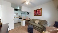 This single-floor unit offers a practical and well-organized open-concept layout, where the living, dining, and kitchen areas flow naturally into one another. The living area is anchored by a comfortable sofa and features carpeted flooring, a ceiling fan with a light fixture overhead, and a textured ceiling. A tufted ottoman serves as a versatile centerpiece, and framed wall art adds a touch of warmth and color to the space. Adjacent to the living area, a compact dining nook features a raised breakfast bar-style counter with wooden chairs, ideal for casual meals or quick bites. The kitchen is thoughtfully designed with white cabinetry, a matching refrigerator, a dishwasher, and a gas range. A range hood is integrated into the cabinetry above the cooktop, and the countertops provide ample workspace for meal preparation. Tile flooring in the kitchen and dining areas transitions smoothly to carpet in the living space. A window in the kitchen allows for natural light to filter in, contributing to the overall brightness of the unit. The layout is efficient and functional, making excellent use of the available space throughout.