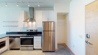 This single-floor unit offers a thoughtfully designed layout that balances modern finishes with functional living. The kitchen is a standout feature, equipped with stainless steel appliances including a refrigerator and electric range, a sleek stainless steel wall-mounted range hood, dark granite countertops, crisp white cabinetry, and a peninsula with additional counter space and a modern faucet. Track lighting illuminates the kitchen, highlighting the open shelving and clean lines throughout. Polished concrete floors run throughout the unit, providing a cool, industrial aesthetic that ties the spaces together cohesively. The bedroom is a bright, comfortable room featuring a window that allows natural light to fill the space, along with a wall-mounted air conditioning unit and a closet with built-in wire shelving for organized storage. The bathroom is well-appointed with a dark granite vanity countertop, white cabinetry, a tub-shower combination, and continued concrete flooring. An additional closet adjacent to the bathroom offers further storage with wire shelving. The unit also includes a dedicated laundry area with a stacked front-loading washer and dryer alongside generous wire shelving, adding significant convenience and practicality to everyday living.