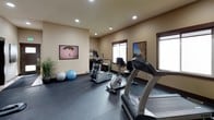 Located at 12202 East Maxwell Avenue in Spokane, Washington, this single-floor fitness space is thoughtfully designed to support a complete workout experience. The room features durable rubber flooring throughout, providing a safe and functional surface for a wide range of exercises.

Cardio equipment is well-represented, with a treadmill, elliptical trainer, and stationary bike positioned to take advantage of the natural light streaming in through multiple windows. Recessed ceiling lighting ensures the space remains bright and inviting at any time of day.

A dedicated free weights area includes a dumbbell rack stocked with a range of weights, complemented by a workout bench for strength training. Stability balls add versatility for core and flexibility exercises. A large wall-mounted mirror helps users monitor their form, while motivational athletic artwork displayed across the walls contributes to an energizing atmosphere.

Entertainment is provided by a wall-mounted flat-screen television, making workouts more engaging. The warm, neutral-toned walls and dark wood-trimmed windows and doors lend the space a polished, cohesive aesthetic. A private entrance door ensures easy, dedicated access to the fitness area. Overall, this space delivers a comprehensive, well-equipped environment suited for cardio, strength, and functional fitness training.