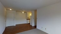This single-floor residential unit offers a clean, functional layout with a seamless flow between the main living area and kitchen. The living space features warm hardwood flooring that transitions to carpet, creating distinct zones within the open floor plan. A ceiling fan is centrally mounted, providing both air circulation and ambient lighting to the room. Crisp white walls throughout lend a bright, neutral backdrop that makes the space feel open and easy to personalize.

A structural column separates the living area from the kitchen, adding architectural definition without fully closing off the two spaces. The kitchen is well-appointed with white upper cabinetry, under-cabinet lighting, and a white refrigerator, giving it a tidy, cohesive appearance. The dark countertop provides a subtle contrast to the light cabinetry. A thermostat is conveniently mounted on the wall near the living area, reflecting practical, user-friendly design throughout.

Overall, the unit presents a straightforward and livable single-level layout, ideal for those seeking a low-maintenance space with a comfortable, functional interior. The combination of hardwood flooring, carpeted areas, and a connected kitchen makes this a versatile and welcoming space.