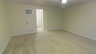 This single-floor space presents a straightforward, functional layout centered around a spacious main room finished with wall-to-wall beige carpeting. The room is painted in a soft, neutral tone that lends a clean and understated feel throughout. A ceiling-mounted light fixture provides overhead illumination, while a pull-cord pendant light offers supplemental lighting options. A floor-level HVAC vent unit is positioned along one wall, ensuring climate control within the space. A wall-mounted electrical outlet and switch plate are also visible, contributing to the room's practical utility. White baseboards trim the perimeter, adding a subtle contrast to the neutral walls. An open doorway leads into a hallway, suggesting additional rooms beyond the main area. The overall ambiance is simple and neutral, offering a blank canvas well-suited for a variety of uses. The space is uncluttered and move-in ready, with its modest, no-frills design making it highly adaptable to different furnishing arrangements and personal styles.