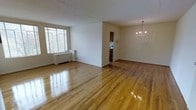 This single-floor space showcases a bright, open layout centered around gleaming hardwood floors that reflect natural light throughout. The main living area is generously proportioned, with white walls and a textured ceiling that lend a clean, neutral backdrop. A large window fitted with horizontal blinds allows abundant natural light to fill the room, complemented by a radiator unit positioned beneath it — a classic heating feature. A partial wall divides the living area from the dining space, which is anchored by an elegant chandelier with candelabra-style bulbs, adding a touch of traditional charm. A passthrough opening in the dividing wall connects to a glimpse of the kitchen beyond, where wood cabinetry is visible. The continuous hardwood flooring flows seamlessly throughout the entire space, enhancing the sense of openness and cohesion. The overall ambiance is understated and classic, offering a versatile, move-in-ready environment that balances functional layout with timeless architectural details.
