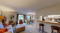This thoughtfully designed single-floor residence offers a comfortable and cohesive layout encompassing a living room, dining room, bedroom, bathroom, hallway, and two closets. The open-concept living and dining area forms the heart of the home, where soft carpeting and neutral walls create a warm, inviting atmosphere. The living room features a tufted sofa and ottoman, accent seating, and a pass-through bar counter that connects to the kitchen, adding both functionality and a social element to the space. The dining room is anchored by a classic chandelier and large sliding glass doors that open to a private balcony overlooking lush, mature trees, flooding the room with natural light and serene outdoor views. The spacious bedroom is finished with calming blue-grey accent walls, plush carpeting, a generous closet, and a window framing peaceful garden views, creating a restful retreat. The full bathroom features a tub-shower combination, a vanity with ample counter space, and tile flooring. A hallway connects the private and shared areas of the home, while two closets provide practical storage throughout. The overall ambiance is calm and residential, with a consistent neutral palette and natural light defining each room.