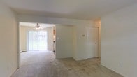 This single-story space features an open layout with neutral-toned carpeting throughout, creating a cohesive and understated aesthetic. The living area flows naturally into an adjoining space, separated by a partial wall that defines each zone while maintaining an open feel. A ceiling fan is positioned in the secondary living or dining area, providing both light and air circulation. Large sliding glass doors flood the space with natural light and offer a direct view of the greenery outside, enhancing the sense of openness. A louvered bifold closet door provides convenient built-in storage along one wall. Visible from the main living area, the kitchen features cabinetry and appliances tucked neatly into its own defined space. The overall palette is light and neutral, with white walls and beige carpeting contributing to a clean, move-in-ready environment. The single-floor layout ensures easy accessibility throughout, with a practical and functional design that suits everyday living.