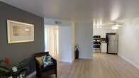 This single-floor space presents a clean, open layout that connects the living area and kitchen in a cohesive flow. Light wood laminate flooring runs throughout, lending warmth and visual continuity across the space. The living area features a dark accent wall in a cool grey tone, creating contrast against the bright white trim and walls. A ceiling-mounted ventilation unit and recessed lighting ensure comfort and functionality throughout.

The kitchen is well-appointed with stainless steel appliances, including a gas range, microwave, and refrigerator. White cabinetry extends to the ceiling, maximizing storage while maintaining a bright, airy feel. Track lighting above the kitchen provides ample task illumination.

A central structural column divides the living space from the hallway, which leads to additional rooms beyond. The hallway benefits from natural light filtering through, keeping the interior feeling open and welcoming. White baseboards and clean wall finishes throughout contribute to a polished, move-in-ready aesthetic. The overall ambiance is modern and functional, with a neutral palette that allows for versatile furnishing and décor options.