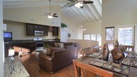This single-floor home makes a striking impression with its dramatic vaulted ceilings accented by exposed white-painted wooden beams, lending the interior an airy, expansive feel. Dual ceiling fans circulate throughout the open-concept main living area, which flows effortlessly between the living room, dining area, and kitchen. The living room is anchored by a generous sectional leather sofa, complemented by rich, warm hardwood floors that run throughout the space. A decorative chandelier adds an elegant touch above the dining area, which features a substantial dark wood table with seating for multiple guests. The kitchen is appointed with dark espresso cabinetry, dark granite countertops, stainless steel appliances including a gas range and microwave, and a sink positioned along the counter. Tall upper walls provide space for decorative greenery, adding a natural element to the interior. Windows along the far wall invite natural light into the dining and living areas, offering views to the exterior. The cohesive design, combining dark wood tones, warm neutral walls, and soaring architectural ceilings, creates an inviting and functional space well-suited for both everyday living and entertaining.