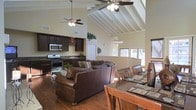 This single-floor home makes a striking impression with its dramatic vaulted ceilings accented by exposed white-painted wooden beams, lending the interior an airy, expansive feel. Dual ceiling fans circulate throughout the open-concept main living area, which flows effortlessly between the living room, dining area, and kitchen. The living room is anchored by a generous sectional leather sofa, complemented by rich, warm hardwood floors that run throughout the space. A decorative chandelier adds an elegant touch above the dining area, which features a substantial dark wood table with seating for multiple guests. The kitchen is appointed with dark espresso cabinetry, dark granite countertops, stainless steel appliances including a gas range and microwave, and a sink positioned along the counter. Tall upper walls provide space for decorative greenery, adding a natural element to the interior. Windows along the far wall invite natural light into the dining and living areas, offering views to the exterior. The cohesive design, combining dark wood tones, warm neutral walls, and soaring architectural ceilings, creates an inviting and functional space well-suited for both everyday living and entertaining.