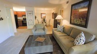 This single-story space offers an open-concept floor plan that seamlessly connects the living room, dining area, and kitchen into one cohesive and functional layout. The living room serves as the central gathering space, featuring plush carpet flooring and warm, neutral-toned walls that create an inviting atmosphere. A generously sized sofa and matching oversized armchair are arranged around a large tufted ottoman, providing a comfortable and well-appointed seating area. Decorative wall art and a tall floor lamp contribute to the room's warm ambiance.

The dining area is positioned adjacent to the living space, anchored by a round glass-top table surrounded by chairs and illuminated by an elegant pendant light fixture overhead. This transitional zone flows naturally into the kitchen, which features wood-toned cabinetry, dark appliances including a range and refrigerator, and a practical layout suited for everyday cooking. The open sightlines between all three areas enhance the sense of spaciousness throughout. Overall, the space presents a cohesive, comfortable, and well-organized interior designed for easy everyday living.