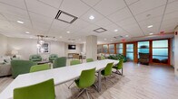 This well-appointed multipurpose space is thoughtfully designed to accommodate a variety of functions, from collaborative meetings to casual social gatherings. The open floor plan is unified by light wood-tone flooring and a suspended tile ceiling with recessed and pendant lighting throughout, creating a bright and welcoming ambiance.

At the heart of the space sits a long, white conference table surrounded by vibrant green upholstered chairs — ideal for group meetings or collaborative work sessions. Adjacent to this is a more intimate café-style area with white tables and modern bar-height seating beneath stylish globe pendant lights.

A dedicated lounge zone features a generous sectional sofa in neutral tones accented with green throw pillows, accompanied by additional green armchairs, providing comfortable areas for relaxed conversation. A wall-mounted flat-screen television anchors one end of the lounge area, set against a textured accent wall.

Two upholstered striped accent chairs create a quieter nook near warm wood-framed frosted glass partitions, which lend privacy and architectural character to the space. A small credenza with a decorative lamp adds a refined touch to this corner.

Throughout the space, curated black-and-white framed artwork contributes a polished, professional aesthetic. Structural columns clad in textured wallcovering serve as subtle design features that define zones without interrupting the open, airy flow of this versatile environment.