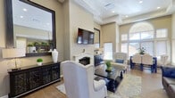 This single-floor community space offers a thoughtfully designed interior that balances comfort and functionality. The main living area serves as the centerpiece of the space, showcasing soaring ceilings, wood-look flooring, and a striking white-mantled fireplace flanked by a wall-mounted flat-screen television. Large framed mirrors amplify the sense of openness, while recessed lighting and natural light from multiple windows create a bright and inviting atmosphere. Elegantly upholstered wingback chairs and tufted seating are arranged around a dark wood coffee table, providing ample seating for gathering and relaxation.

Built-in shelving units line the walls of the adjoining lounge area, displaying decorative accents and greenery, and a wide doorway connects to a naturally lit rotunda-style entry with additional seating. The dining area features a substantial dark wood table surrounded by upholstered chairs and benefits from dramatic arched transom windows that flood the room with natural light, with a glass door providing direct outdoor access.

The kitchen is well-appointed with rich dark wood cabinetry, granite countertops, stainless steel appliances including an electric range and refrigerator, and a microwave, making it fully functional for event use or everyday needs. Lush greenery displayed atop the cabinetry adds a warm, welcoming finishing touch throughout.