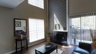 This two-floor space makes an immediate impression with its dramatic double-height ceilings in the living area, creating a grand sense of openness and volume. The focal point of the room is a bold black brick fireplace, topped with a crisp white mantel that provides a clean contrast against the dark surround. Rising behind it, a diagonally laid shiplap accent wall in a weathered gray finish adds texture and architectural character, extending upward to the full height of the vaulted ceiling.

Natural light flows generously into the space through multiple large windows dressed with white horizontal blinds, casting bright, even light across the hardwood-style flooring. A sliding glass door or window on the right side hints at outdoor access, further enhancing the airy feel of the interior.

The living area is thoughtfully arranged with dark-toned furniture, including a substantial coffee table and coordinating seating, anchored by a light area rug. A large floor mirror on the left wall amplifies the sense of space. The neutral warm-toned walls complement the overall palette, resulting in a cohesive, contemporary aesthetic that balances comfort with architectural distinction.