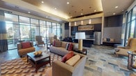 This community room is a thoughtfully designed, single-floor shared space that seamlessly blends comfort and functionality for residents. The open-plan layout is anchored by a generously sized lounge area furnished with neutral-toned sofas and accent chairs accented by rich red and earth-toned throw pillows, arranged to encourage conversation and relaxation. A bold chevron-patterned area rug adds visual interest to the slate tile flooring, which extends throughout the space.

One wall features a built-in dark-wood entertainment center with integrated shelving and a mounted flat-screen television, providing a dedicated media focal point. Recessed lighting and tray ceiling details with warm ambient illumination add architectural depth overhead.

Floor-to-ceiling glass walls and large windows flood the interior with natural light and offer clear views of the landscaped outdoor courtyard, creating a seamless indoor-outdoor connection. Striped wingback chairs positioned near the glazed wall provide a quiet retreat with garden views.

The fully equipped kitchen area showcases dark espresso cabinetry, a mosaic tile backsplash, granite countertops, stainless steel appliances including a refrigerator, range, and microwave, and a large island — making it well-suited for catered events or community gatherings. The overall ambiance is warm, polished, and welcoming.