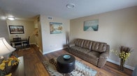 This single-floor space offers a welcoming and functional layout centered around a comfortable living area. The living room features rich, dark hardwood-style flooring that contrasts cleanly against the neutral beige walls, creating a warm and grounded atmosphere. A textured ceiling adds subtle dimension to the space overhead.

The living area is furnished with a tufted brown leather sofa paired with a round dark ottoman serving as a coffee table, anchored by a patterned area rug. Two coastal-themed canvas prints adorn the walls, contributing a relaxed, casual aesthetic to the room. A decorative branch arrangement in the corner adds a touch of natural styling.

A HVAC vent is visible on the wall, and a floor vent ensures efficient climate control throughout the space. The layout flows naturally toward an adjoining area — visible in the background — that appears to connect to a kitchen or hallway, suggesting a practical open-concept arrangement. White baseboards and clean wall transitions between rooms give the interior a tidy, well-maintained appearance. Overall, the space conveys a modest yet comfortable living environment with straightforward, functional design.