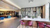 Located at 2287 Welton St in Denver's historic Five Points neighborhood, The Dorsey presents a striking fusion of industrial heritage and contemporary elegance. The entry welcomes visitors with herringbone wood-patterned flooring, exposed brick walls, and floor-to-ceiling glass partitions framed in sleek black metal, setting a refined tone from the moment of arrival.

The reception area is anchored by a sculptural feature wall of layered vertical metallic panels in gold and grey tones, behind which a white quartz-topped desk is paired with plush magenta accent chairs. Natural light pours through expansive windows, illuminating the open-plan layout that connects seamlessly to various functional zones.

A standout communal kitchen and lounge area features deep navy cabinetry set beneath a series of dramatic brick arches, complemented by a boldly tiled island with a white countertop and suspended circular gold pendant lights. Adjacent lounge and co-working areas offer a relaxed yet productive atmosphere, with glass-enclosed private offices providing visual connectivity while maintaining acoustic separation.

Throughout the space, rich textures — including geometric slate tile, warm herringbone wood, and original exposed brick — create a layered aesthetic that balances industrial character with modern sophistication. Lush greenery and curated artwork add warmth, making The Dorsey a vibrant and inviting destination.