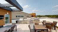 This single-floor space combines functional interior areas with an impressive rooftop outdoor amenity. The rooftop terrace is a standout feature, offering an expansive open-air setting complete with a built-in stainless steel grill station that spans a generous length, accommodating multiple grilling units side by side. The terrace is finished with clean light-colored pavers and furnished with wooden dining tables and chairs, making it well-suited for outdoor gatherings. Surrounding rooftop views include neighboring structures and a backdrop of open sky and distant tree lines.

Inside, the bathroom is finished in warm neutral tones with cream-colored walls throughout. It includes a single-sink vanity topped with a white countertop and housed in a wood-toned cabinet with drawers below. A framed rectangular mirror with a weathered wood surround is mounted above the vanity. The toilet area is tucked into a dedicated alcove, with a small wooden wall-mounted rack positioned above. Brass-toned hardware and outlet covers appear consistently throughout the interior, lending a cohesive, classic finish. The overall interior ambiance is understated and functional, while the rooftop terrace provides a notable outdoor extension of the space.