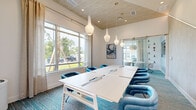 Located in Panama City Beach, Florida, this well-appointed meeting space offers two distinct rooms designed for professional gatherings and collaborative sessions. The main conference room is anchored by a long, segmented white table surrounded by plush teal velvet chairs, creating a vibrant yet refined atmosphere. Elevated ceilings with a warm wood-paneled finish lend an open, airy feel, while a trio of elegant pendant lights in a teardrop silhouette provide focused illumination. Large windows along one wall flood the room with natural light and offer views of lush greenery and palm trees outside. A diagonal light rail accent on the textured wall adds a modern architectural touch, and a framed abstract artwork completes the room's coastal aesthetic. The teal-patterned carpet ties the coastal color palette together throughout both rooms. The secondary room features a natural wood table paired with cream upholstered chairs, bathed in light from floor-to-ceiling windows draped with soft linen curtains. A striking gold leaf-motif chandelier serves as the focal point overhead. A sliding glass partition connects both spaces, offering flexibility for open-plan use or private sessions. Together, these rooms deliver a professional, design-forward environment ideal for meetings, presentations, or collaborative work.