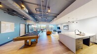 This stylish single-floor amenity space combines industrial design elements with contemporary finishes to create a dynamic and inviting environment. The open-plan layout is anchored by exposed ductwork and structural beams painted in a moody charcoal tone, contrasting beautifully against light hardwood flooring that runs throughout the entire space.

At the heart of the room sits a full-size pool table, complete with a mounted cue rack on the wall, making it the focal point of the entertainment area. Adjacent to it, a comfortable lounge zone features a rich caramel leather sofa, coordinating blue accent chairs, and a flat-screen television mounted on the wall, all bathed in natural light from expansive floor-to-ceiling windows and sliding glass doors that open to an outdoor terrace with additional seating.

The kitchen area is well-appointed, showcasing a generous white quartz island with a bold cobalt blue base and seating for multiple guests. Pendant lighting illuminates the island, while slate-blue cabinetry, a white subway tile backsplash, open shelving, and stainless steel appliances complete the modern culinary setup. A casual high-top dining table near the terrace access adds further flexibility for gathering and socializing. This space is thoughtfully designed for community and entertainment.